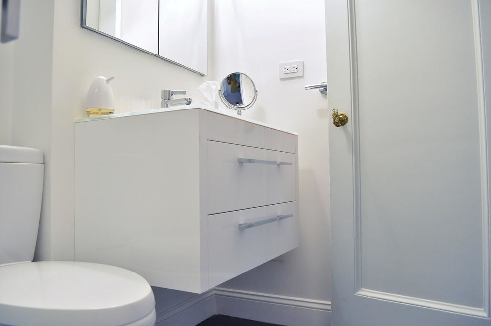 Inspiration for a small modern 3/4 bathroom in New York with a wall-mount sink, flat-panel cabinets, white cabinets, glass benchtops, a corner shower, subway tile, white walls, a two-piece toilet, white tile and vinyl floors.