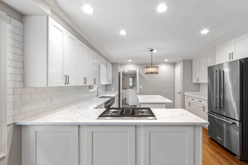 Inspiration for a large coastal l-shaped medium tone wood floor, brown floor and tray ceiling eat-in kitchen remodel in DC Metro with a farmhouse sink, shaker cabinets, white cabinets, quartz countertops, gray backsplash, ceramic backsplash, stainless steel appliances, an island and white countertops