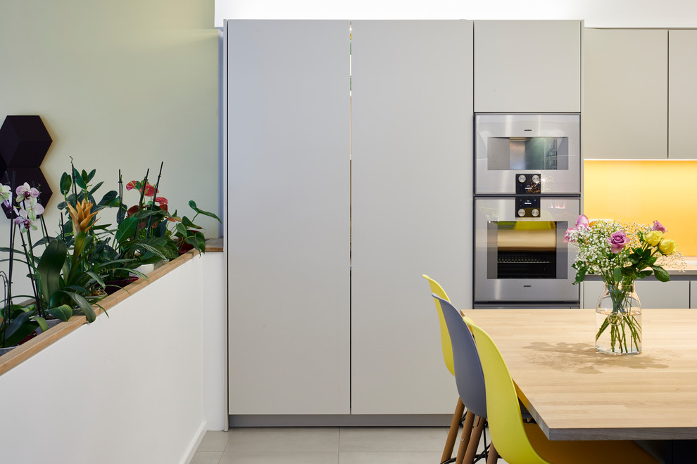 Large modern single-wall open plan kitchen with black cabinets, stainless steel benchtops, yellow splashback, glass sheet splashback, stainless steel appliances, ceramic floors, with island and grey floor.