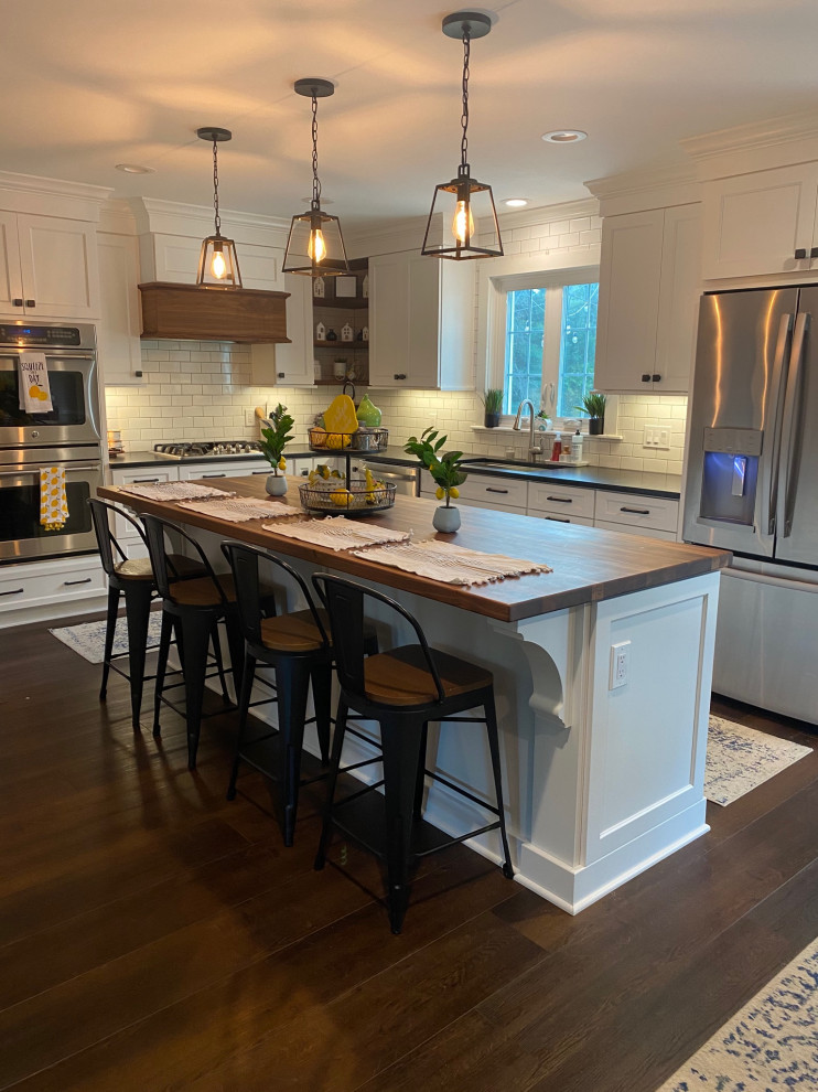 Photo of a large country l-shaped eat-in kitchen in Other with an undermount sink, recessed-panel cabinets, white cabinets, wood benchtops, white splashback, ceramic splashback, stainless steel appliances, dark hardwood floors, with island and brown floor.