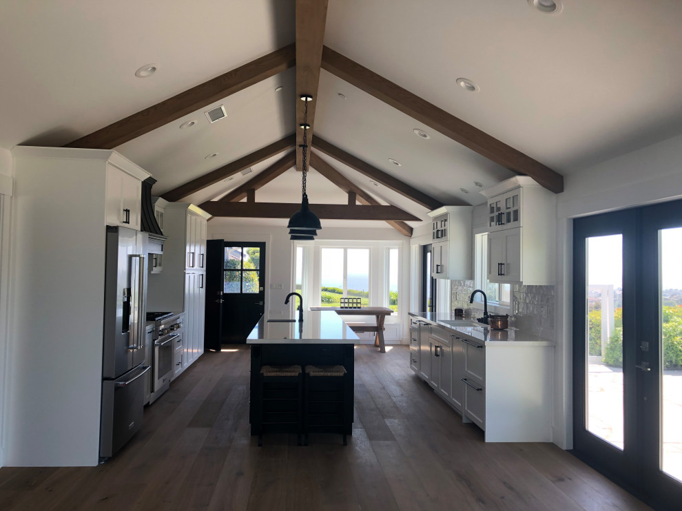 This is an example of a mid-sized country galley open plan kitchen in Orange County with an undermount sink, shaker cabinets, white cabinets, quartz benchtops, white splashback, ceramic splashback, stainless steel appliances, medium hardwood floors, with island, white benchtop and vaulted.