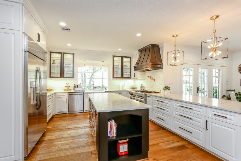Enclosed kitchen - large transitional u-shaped medium tone wood floor and brown floor enclosed kitchen idea in Houston with a farmhouse sink, raised-panel cabinets, white cabinets, granite countertops, white backsplash, glass tile backsplash, stainless steel appliances, an island and white countertops
