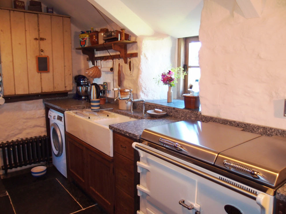 Small rustic u-shaped enclosed kitchen in Cornwall with a belfast sink, shaker cabinets, dark wood cabinets, granite worktops, white appliances, slate flooring, no island, grey floors, grey worktops and exposed beams.