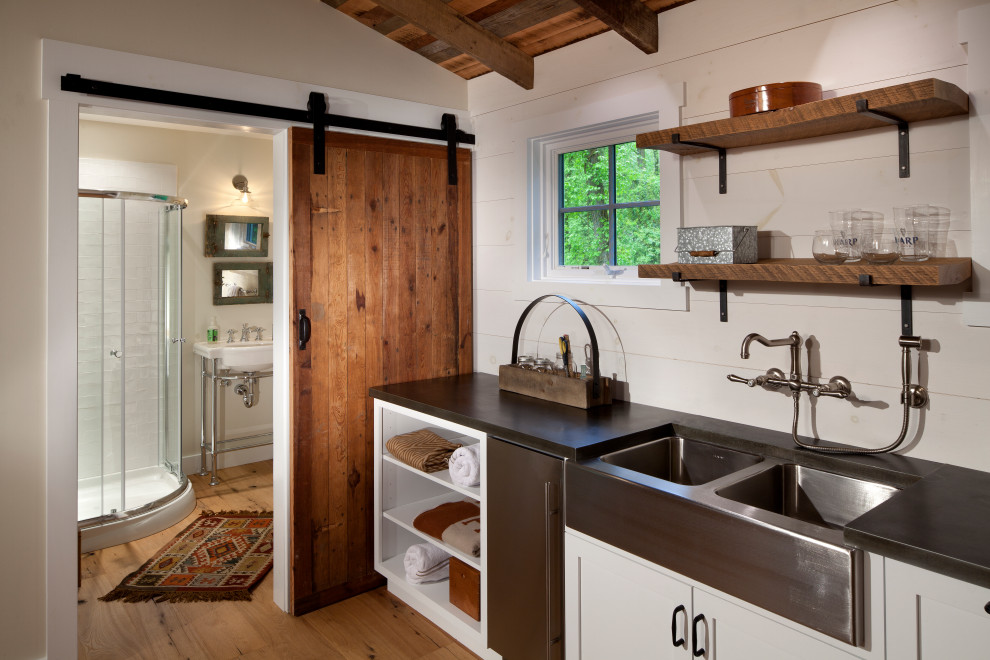 This is an example of a country single-wall open plan kitchen in Baltimore with a double-bowl sink, shaker cabinets, white cabinets, white splashback, timber splashback, stainless steel appliances, medium hardwood floors, no island, brown floor, black benchtop and exposed beam.