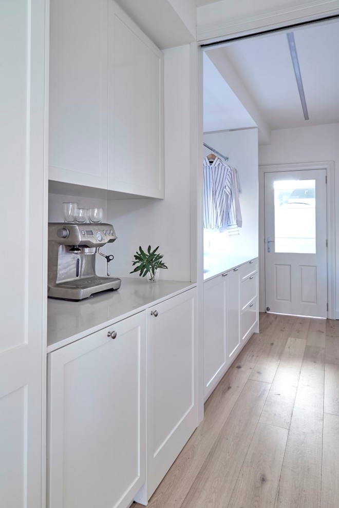 Inspiration for a mid-sized transitional galley utility room in Sydney with an undermount sink, shaker cabinets, quartz benchtops, white walls, light hardwood floors, a concealed washer and dryer and white cabinets.