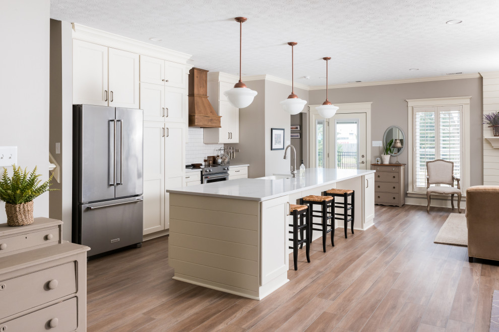 Photo of a mid-sized transitional open plan kitchen in Indianapolis with shaker cabinets, white cabinets, quartz benchtops, white splashback, stainless steel appliances, medium hardwood floors, with island, grey benchtop, a farmhouse sink and brown floor.