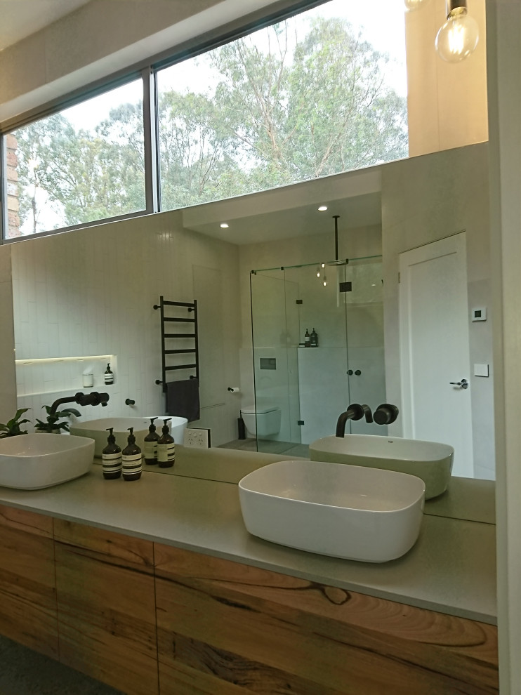 Large modern bathroom in Melbourne with flat-panel cabinets, medium wood cabinets, a freestanding bath, a one-piece toilet, white tiles, ceramic tiles, white walls, terrazzo flooring, a vessel sink, engineered stone worktops, grey floors, a hinged door, a wall niche, double sinks, a floating vanity unit, a vaulted ceiling and grey worktops.