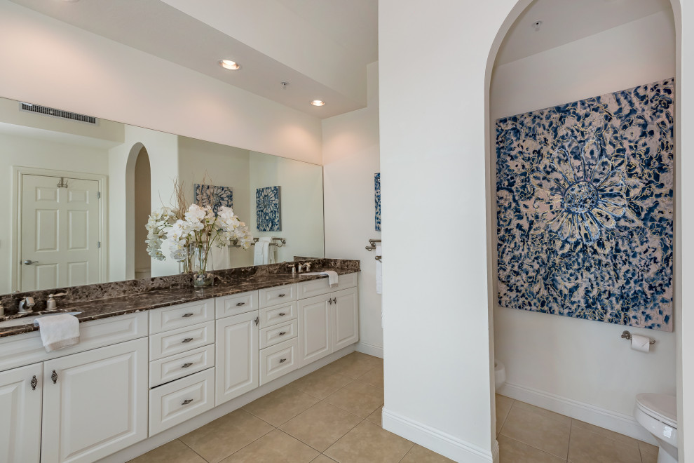 Photo of a large modern master bathroom in Tampa with raised-panel cabinets, beige cabinets, a drop-in tub, a corner shower, a two-piece toilet, beige tile, travertine, white walls, travertine floors, an undermount sink, granite benchtops, beige floor, a hinged shower door and brown benchtops.