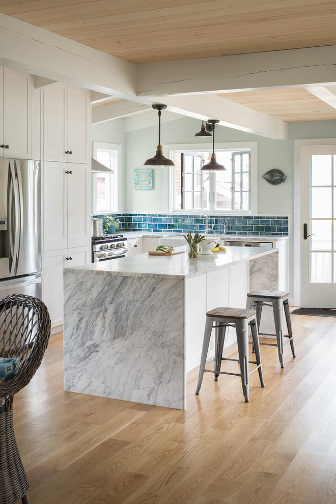 Photo of a beach style l-shaped kitchen in Portland Maine with a farmhouse sink, shaker cabinets, white cabinets, blue splashback, subway tile splashback, stainless steel appliances, light hardwood floors, with island and multi-coloured benchtop.