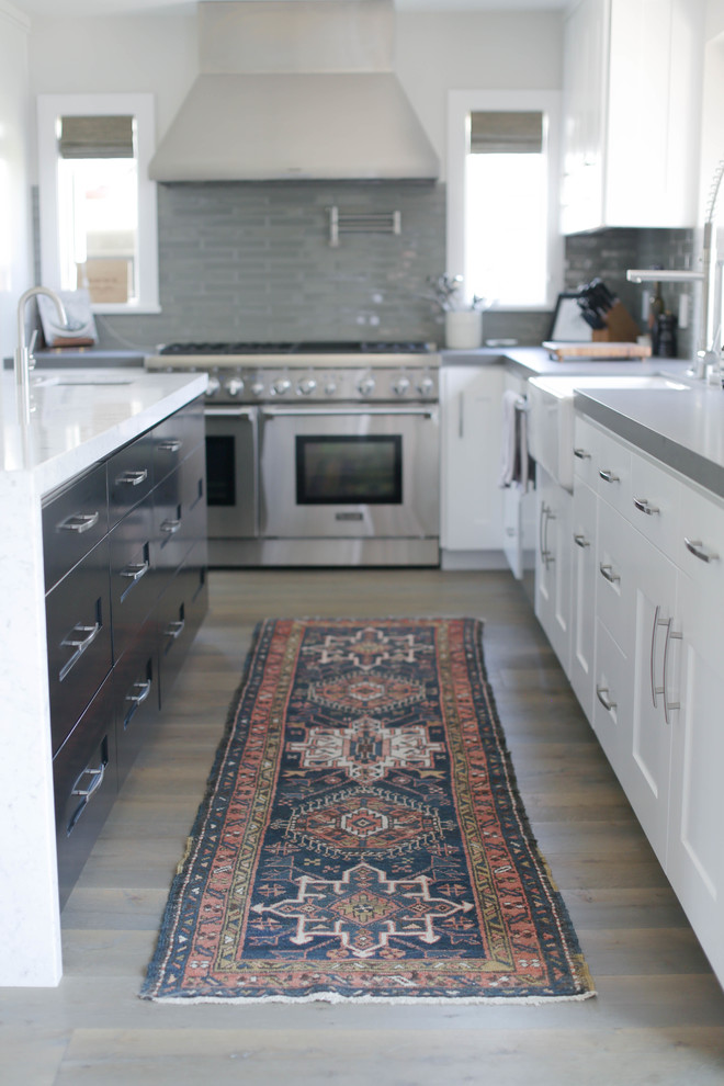 Open concept kitchen - large transitional medium tone wood floor and brown floor open concept kitchen idea in Orange County with a farmhouse sink, shaker cabinets, white cabinets, quartzite countertops, gray backsplash, glass tile backsplash, stainless steel appliances and an island