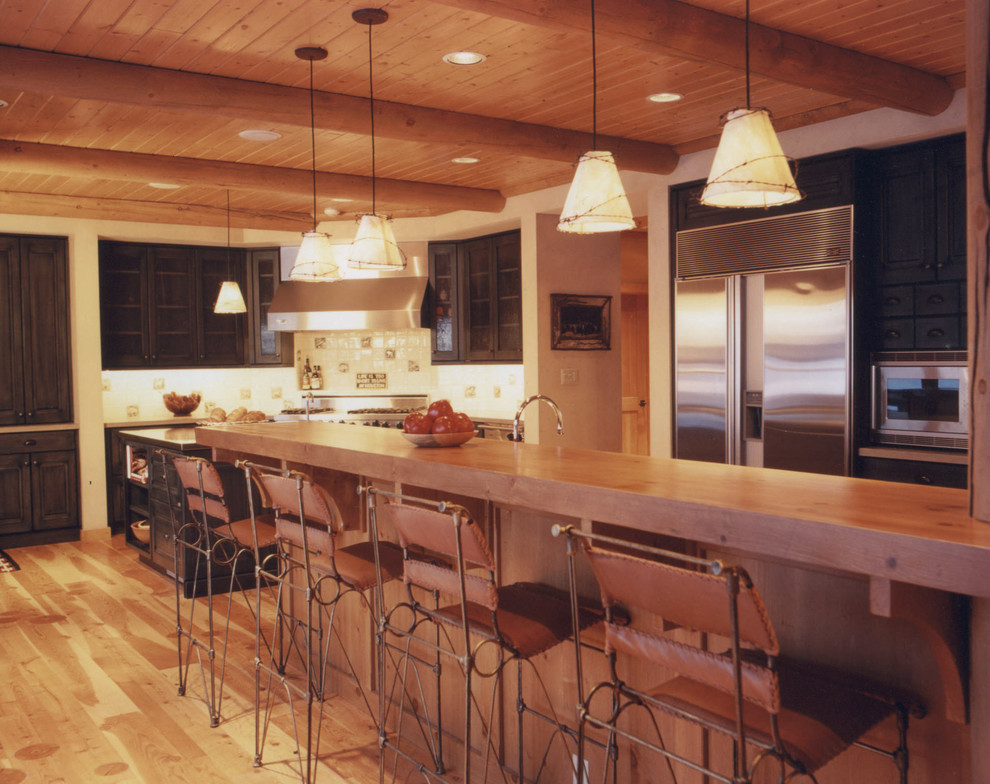 Large mountain style u-shaped light wood floor eat-in kitchen photo with glass-front cabinets, dark wood cabinets, wood countertops, white backsplash, porcelain backsplash, stainless steel appliances and an island