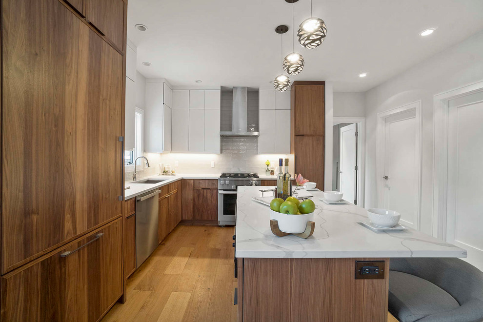 Photo of a mid-sized modern l-shaped eat-in kitchen in Boston with a drop-in sink, flat-panel cabinets, brown cabinets, quartzite benchtops, white splashback, ceramic splashback, stainless steel appliances, medium hardwood floors, with island, brown floor and white benchtop.