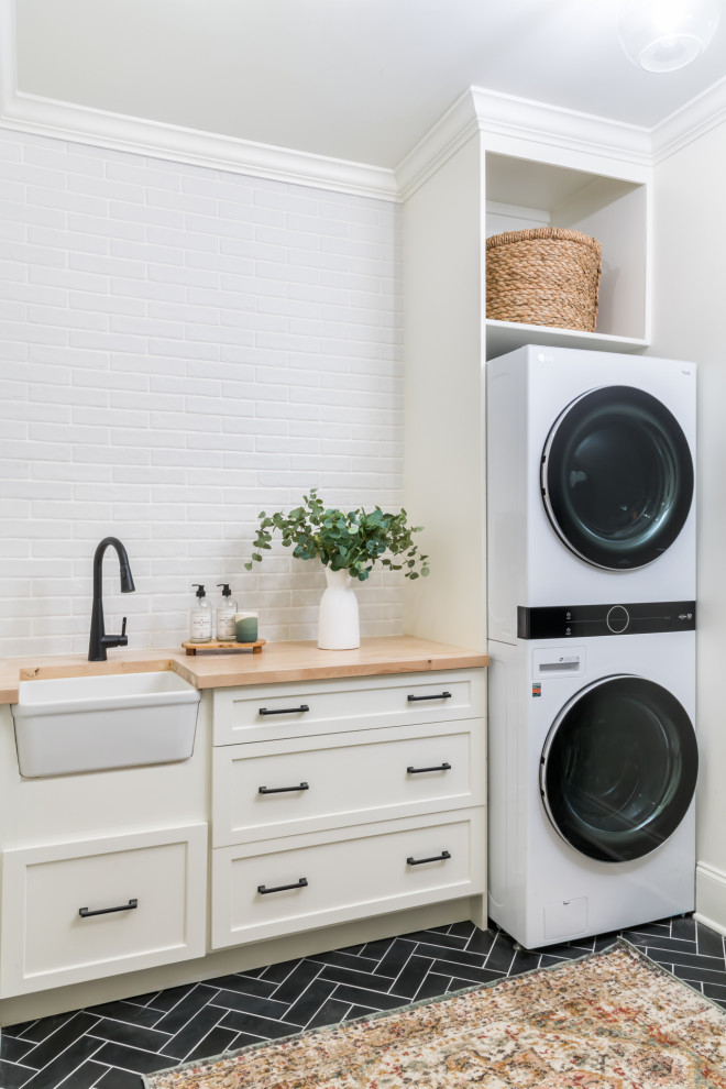 Design ideas for a medium sized traditional single-wall utility room in Atlanta with a belfast sink, raised-panel cabinets, white cabinets, wood worktops, white splashback, brick splashback, white walls, ceramic flooring, a stacked washer and dryer and black floors.