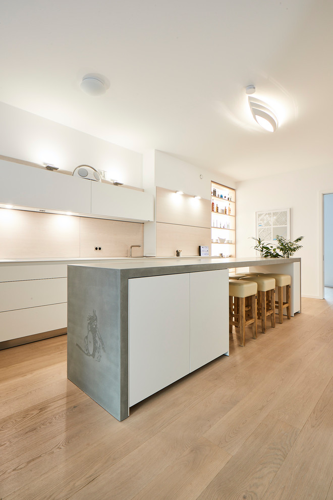 This is an example of a large modern galley open plan kitchen in Hamburg with an integrated sink, flat-panel cabinets, white cabinets, concrete benchtops, beige splashback, timber splashback, white appliances, medium hardwood floors, with island, brown floor and grey benchtop.