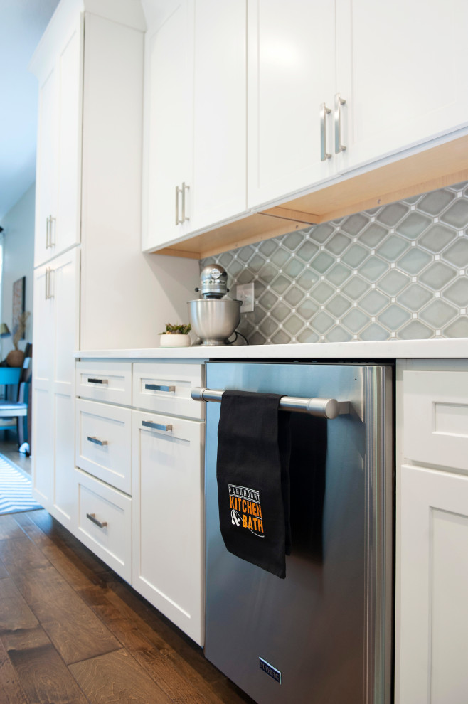 Mid-sized transitional l-shaped medium tone wood floor and brown floor eat-in kitchen photo with an undermount sink, shaker cabinets, white cabinets, quartz countertops, gray backsplash, mosaic tile backsplash, stainless steel appliances, an island and white countertops