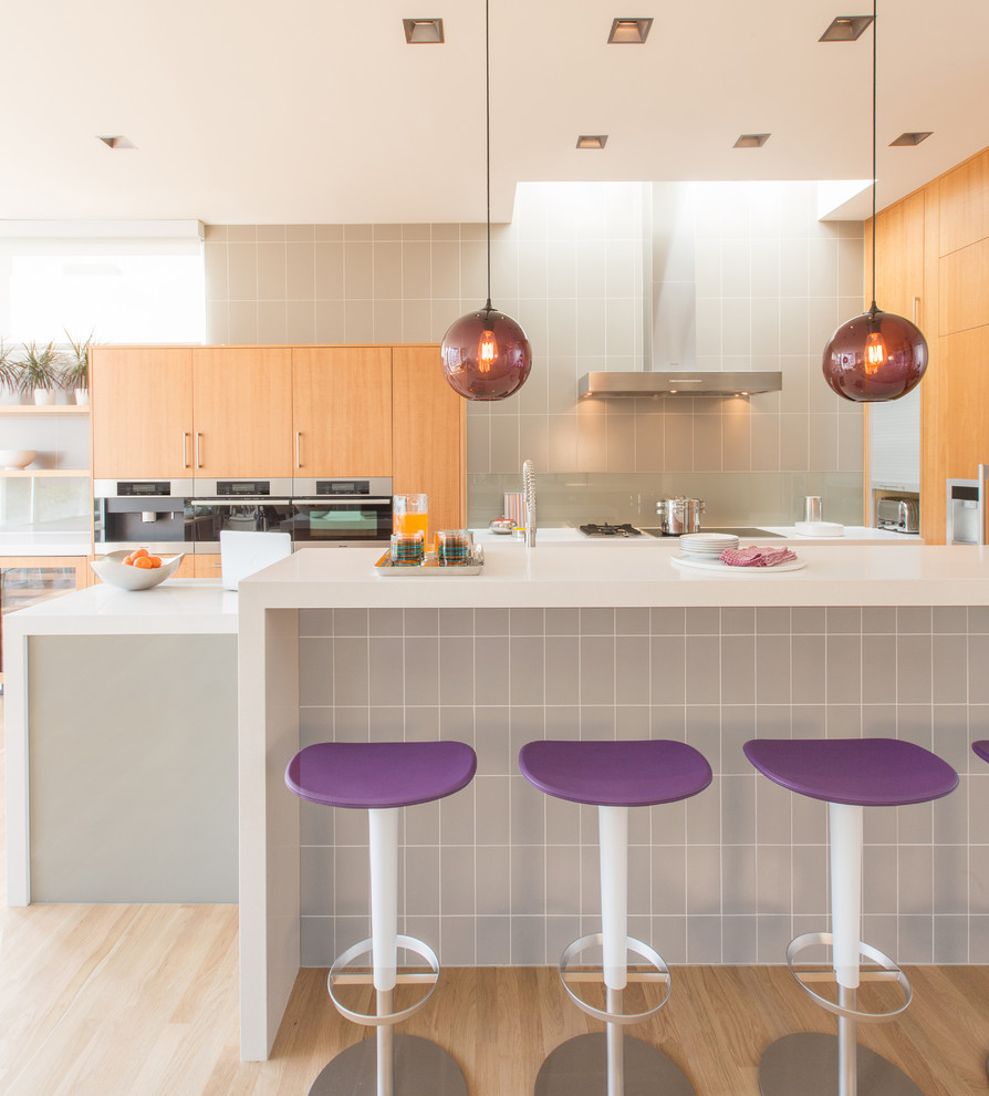 This is an example of a mid-sized contemporary galley eat-in kitchen in San Francisco with flat-panel cabinets, light wood cabinets, grey splashback, stainless steel appliances, light hardwood floors, quartz benchtops, glass sheet splashback and a peninsula.