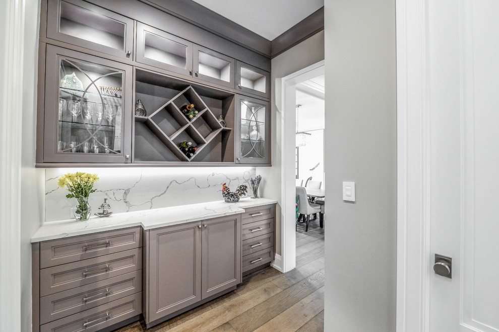 This is an example of a mid-sized transitional single-wall wet bar in New York with recessed-panel cabinets, grey cabinets, marble benchtops, white splashback, marble splashback, medium hardwood floors, brown floor and white benchtop.