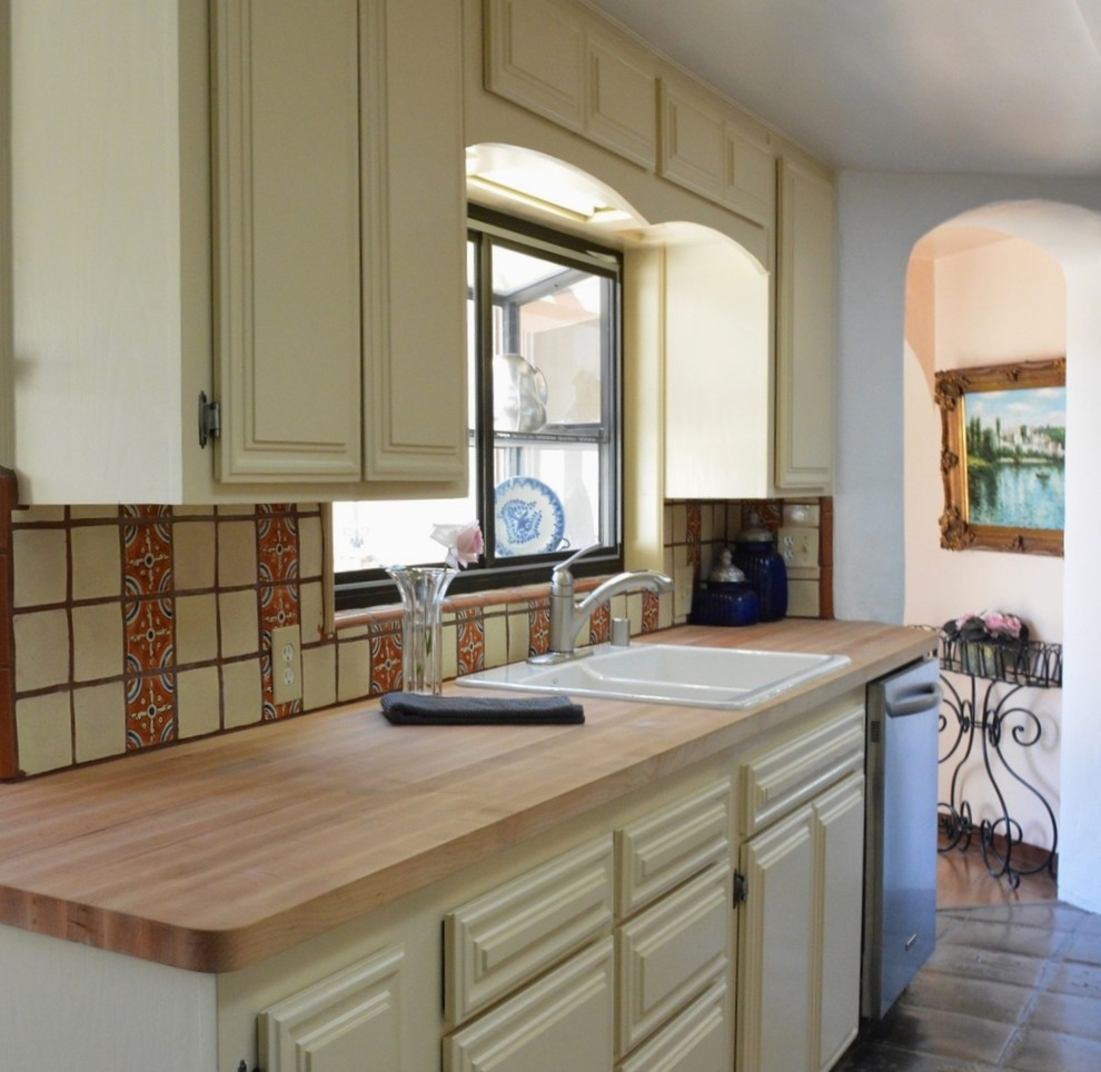 This is an example of a mid-sized mediterranean kitchen in San Diego with a drop-in sink, raised-panel cabinets, wood benchtops, red splashback, ceramic splashback, stainless steel appliances and terra-cotta floors.
