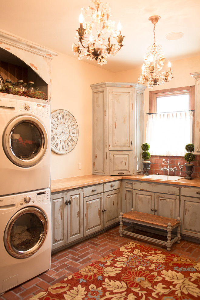 Photo of a mid-sized country l-shaped dedicated laundry room in Oklahoma City with a drop-in sink, raised-panel cabinets, distressed cabinets, wood benchtops, beige walls, brick floors, a stacked washer and dryer, red floor and beige benchtop.