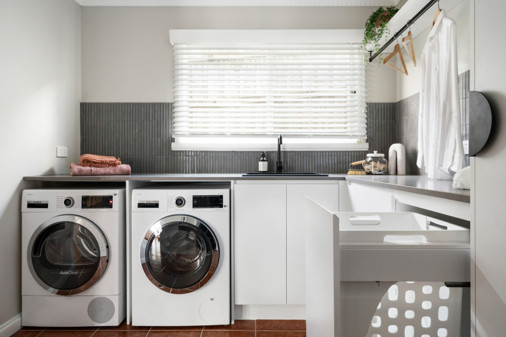 Photo of a mid-sized contemporary l-shaped utility room in Melbourne with a drop-in sink, flat-panel cabinets, white cabinets, quartz benchtops, grey splashback, matchstick tile splashback, white walls, porcelain floors, a side-by-side washer and dryer, orange floor and grey benchtop.
