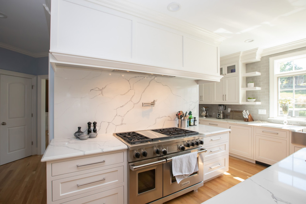Photo of a mid-sized modern u-shaped eat-in kitchen in Boston with a farmhouse sink, beaded inset cabinets, white cabinets, quartz benchtops, grey splashback, glass tile splashback, stainless steel appliances, light hardwood floors, with island, brown floor and white benchtop.