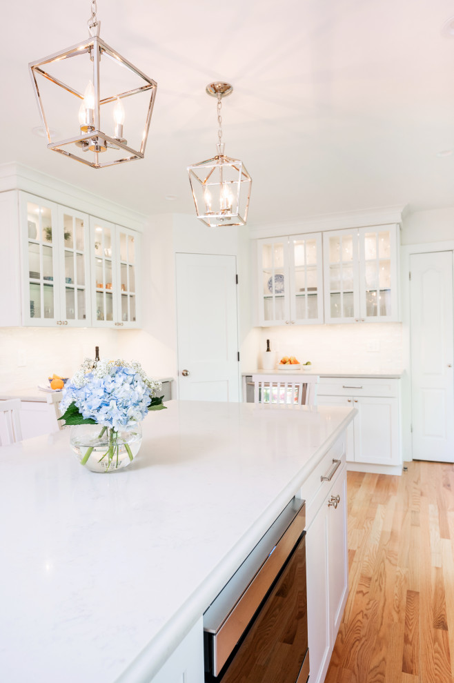 Example of a large transitional l-shaped light wood floor and brown floor kitchen pantry design in Baltimore with an undermount sink, shaker cabinets, white cabinets, quartz countertops, white backsplash, ceramic backsplash, stainless steel appliances, an island and white countertops