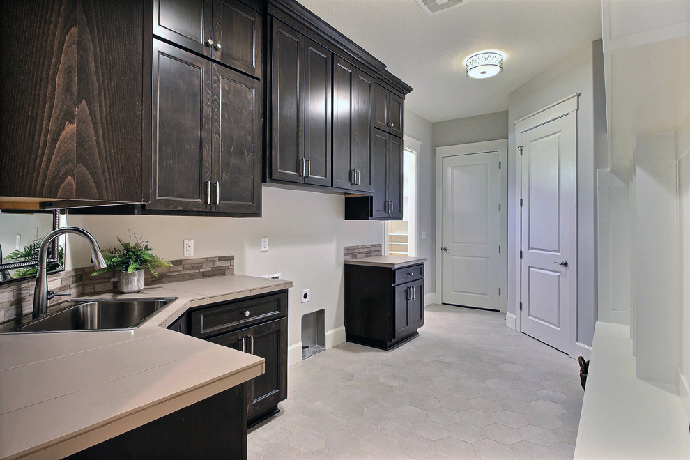 Photo of an expansive transitional galley utility room in Portland with a drop-in sink, recessed-panel cabinets, dark wood cabinets, tile benchtops, white walls, ceramic floors, a side-by-side washer and dryer and grey floor.