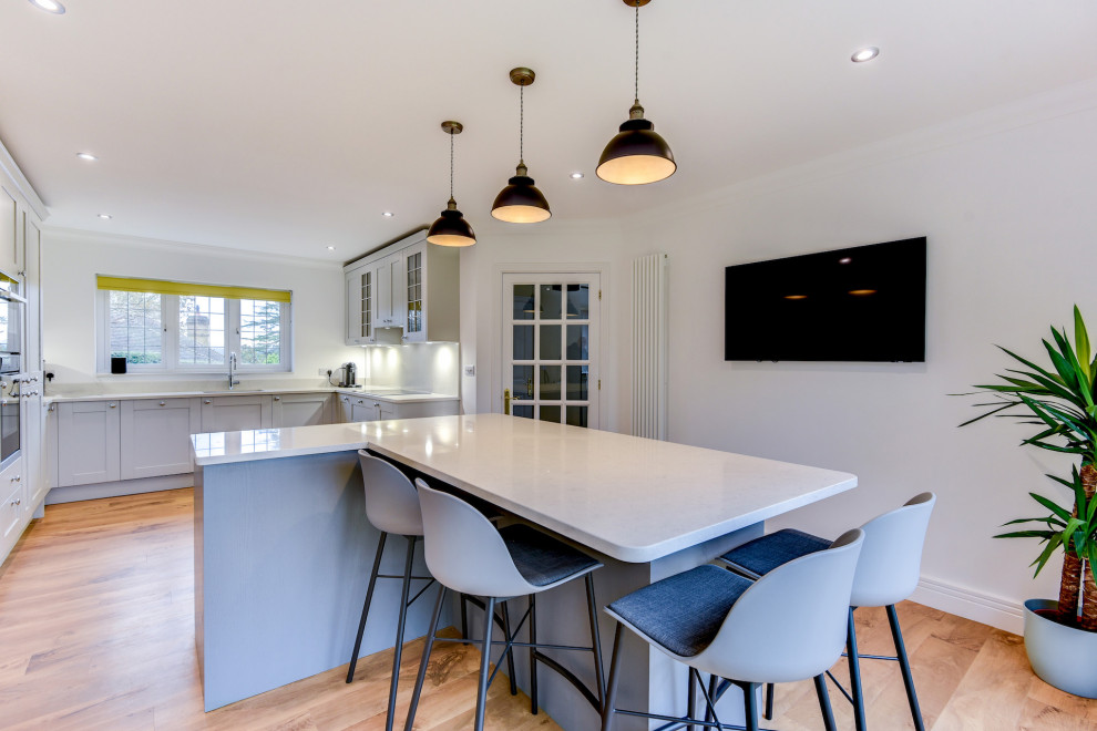 Design ideas for a large contemporary grey and white u-shaped open plan kitchen in Sussex with a submerged sink, shaker cabinets, grey cabinets, quartz worktops, white splashback, integrated appliances, medium hardwood flooring, an island, brown floors, white worktops and feature lighting.