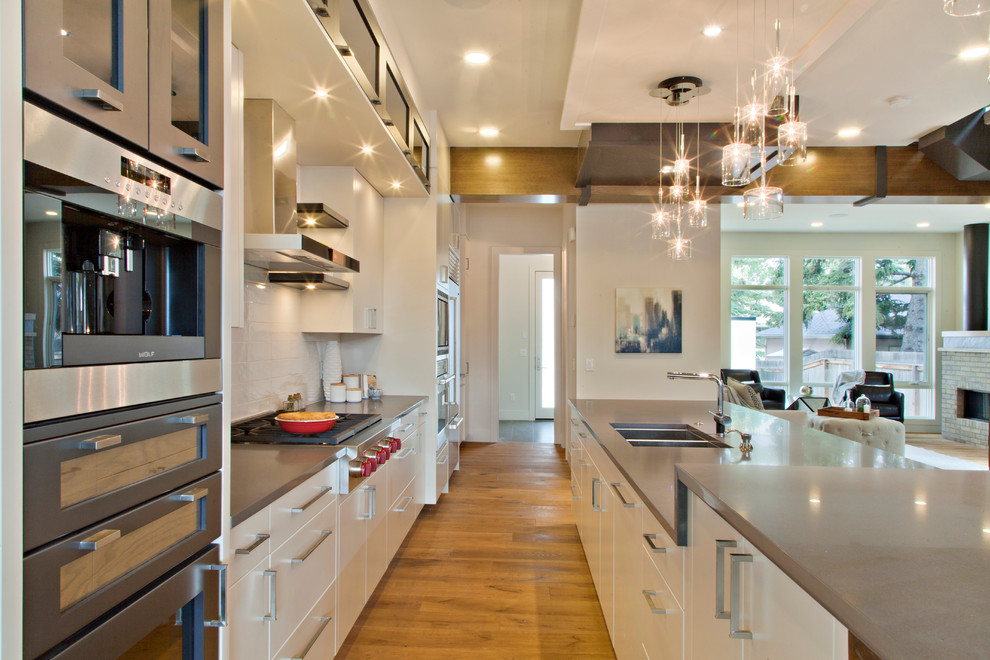 Example of a transitional galley medium tone wood floor open concept kitchen design in Calgary with a double-bowl sink, flat-panel cabinets, white cabinets, solid surface countertops, white backsplash, ceramic backsplash and an island