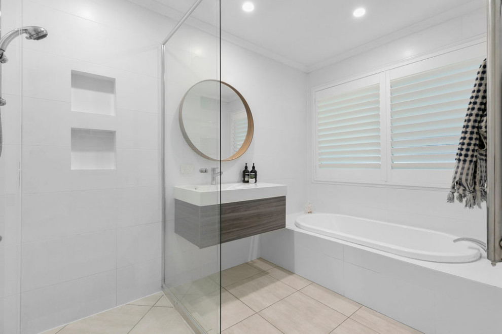 This is an example of a beach style bathroom in Sunshine Coast with furniture-like cabinets, medium wood cabinets, a drop-in tub, a one-piece toilet, white tile, ceramic tile, white walls, porcelain floors, an integrated sink, engineered quartz benchtops, multi-coloured floor, a hinged shower door and white benchtops.