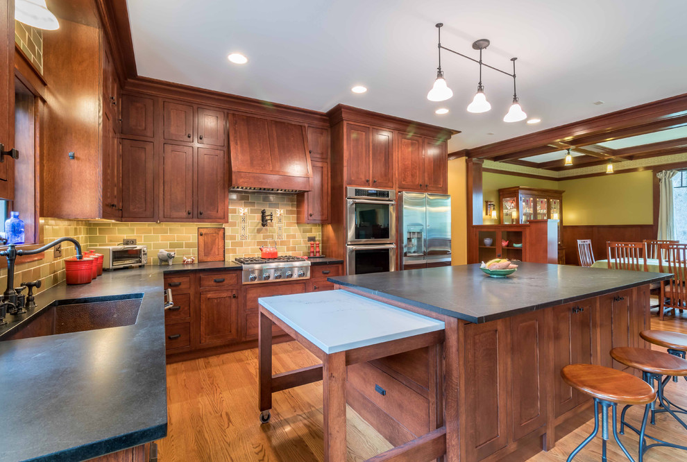 Photo of a mid-sized arts and crafts l-shaped open plan kitchen in Chicago with an undermount sink, shaker cabinets, medium wood cabinets, soapstone benchtops, green splashback, ceramic splashback, stainless steel appliances, light hardwood floors, with island, black benchtop, brown floor and wallpaper.