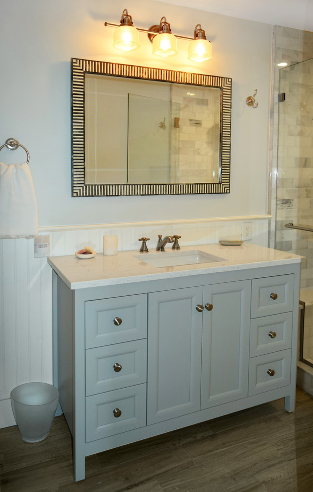 This is an example of a mid-sized transitional master bathroom in Philadelphia with recessed-panel cabinets, grey cabinets, an alcove tub, an alcove shower, a one-piece toilet, gray tile, marble, blue walls, porcelain floors, an undermount sink, engineered quartz benchtops, brown floor, a hinged shower door and white benchtops.