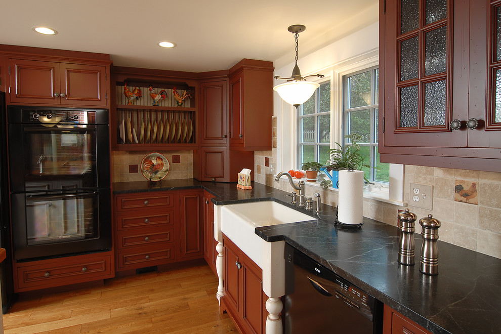 Inspiration for a large traditional l-shaped eat-in kitchen in Boston with a farmhouse sink, recessed-panel cabinets, red cabinets, soapstone benchtops, beige splashback, ceramic splashback, black appliances and medium hardwood floors.