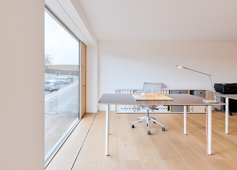 Modern home office in Frankfurt with white walls, light hardwood floors, a freestanding desk and beige floor.