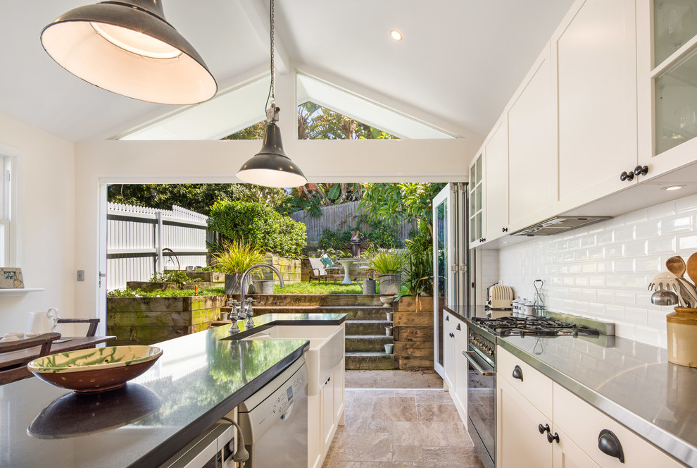 Photo of a mid-sized transitional galley eat-in kitchen in Sydney with glass-front cabinets, white cabinets, stainless steel benchtops, white splashback, ceramic splashback, ceramic floors, with island, a farmhouse sink and black appliances.