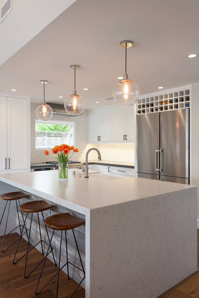 Photo of a contemporary l-shaped eat-in kitchen in Orange County with a farmhouse sink, shaker cabinets, white cabinets, quartz benchtops, white splashback, ceramic splashback and stainless steel appliances.