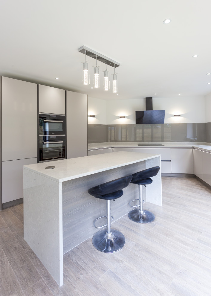 Mid-sized contemporary u-shaped eat-in kitchen in Manchester with an undermount sink, flat-panel cabinets, grey cabinets, quartzite benchtops, brown splashback, glass sheet splashback, black appliances, medium hardwood floors, with island, brown floor and grey benchtop.