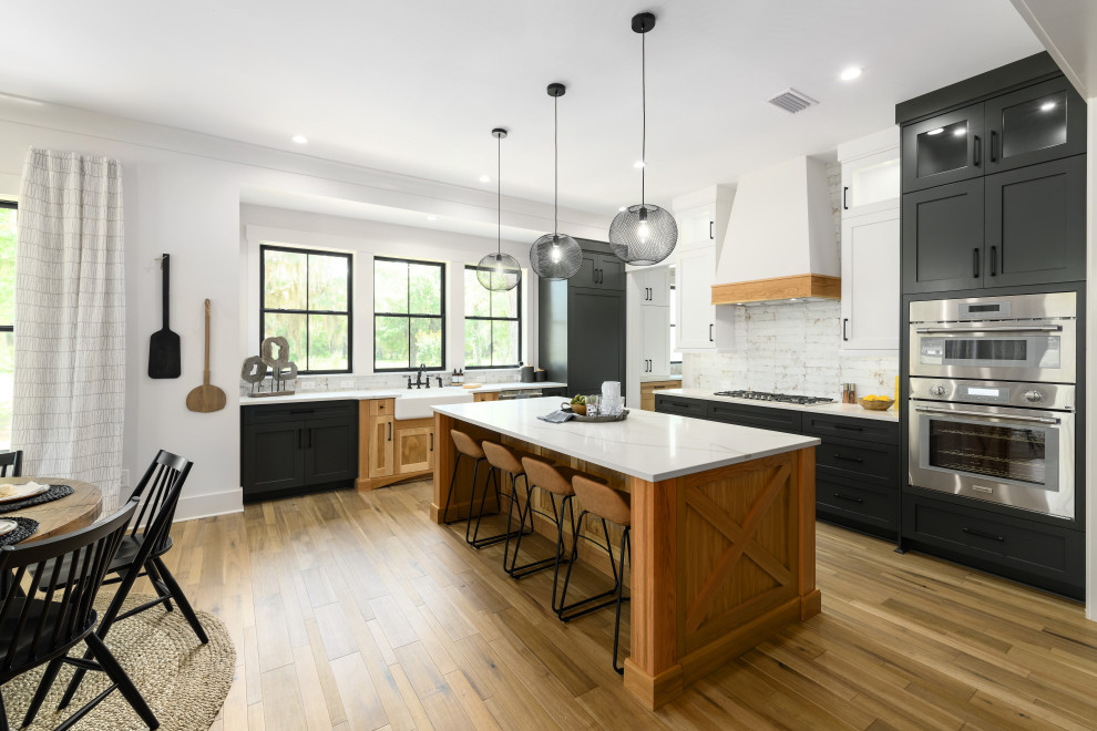 Inspiration for a mid-sized farmhouse u-shaped light wood floor and brown floor eat-in kitchen remodel in Jacksonville with a farmhouse sink, quartz countertops, white backsplash, ceramic backsplash, stainless steel appliances, an island, white countertops, shaker cabinets and black cabinets