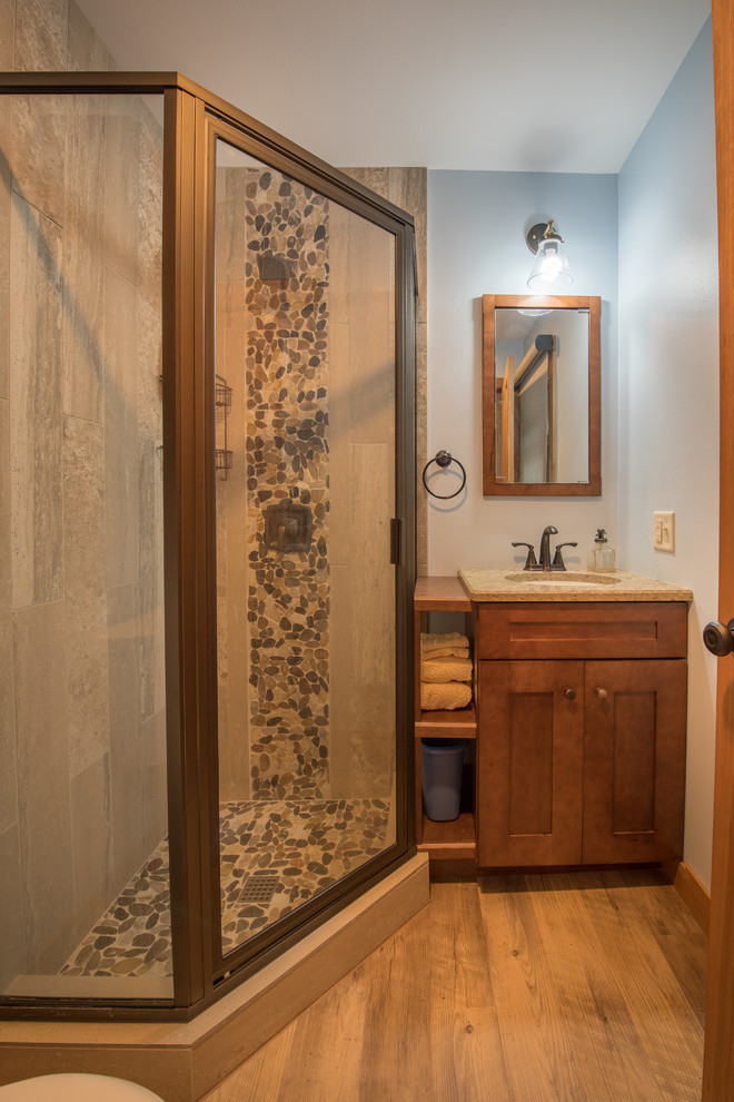 This is an example of a mid-sized arts and crafts 3/4 bathroom in Minneapolis with shaker cabinets, dark wood cabinets, a corner shower, beige tile, pebble tile, blue walls, medium hardwood floors, an undermount sink, granite benchtops, brown floor, a hinged shower door and brown benchtops.