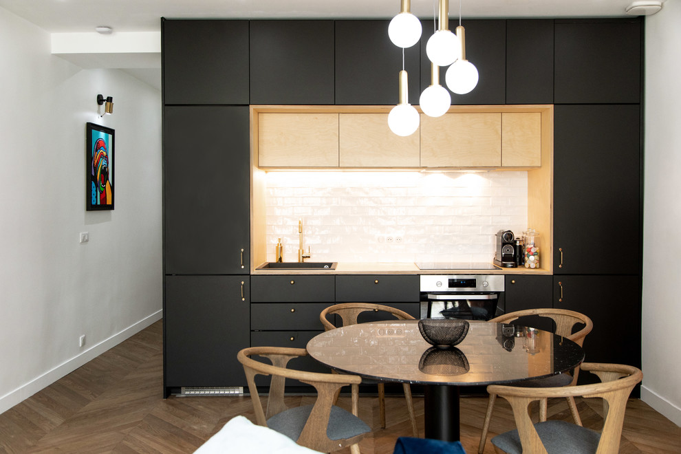 Photo of a small modern single-wall open plan kitchen in Other with flat-panel cabinets, black cabinets, wood benchtops, white splashback, subway tile splashback, black appliances, medium hardwood floors, brown floor, a drop-in sink, no island and beige benchtop.