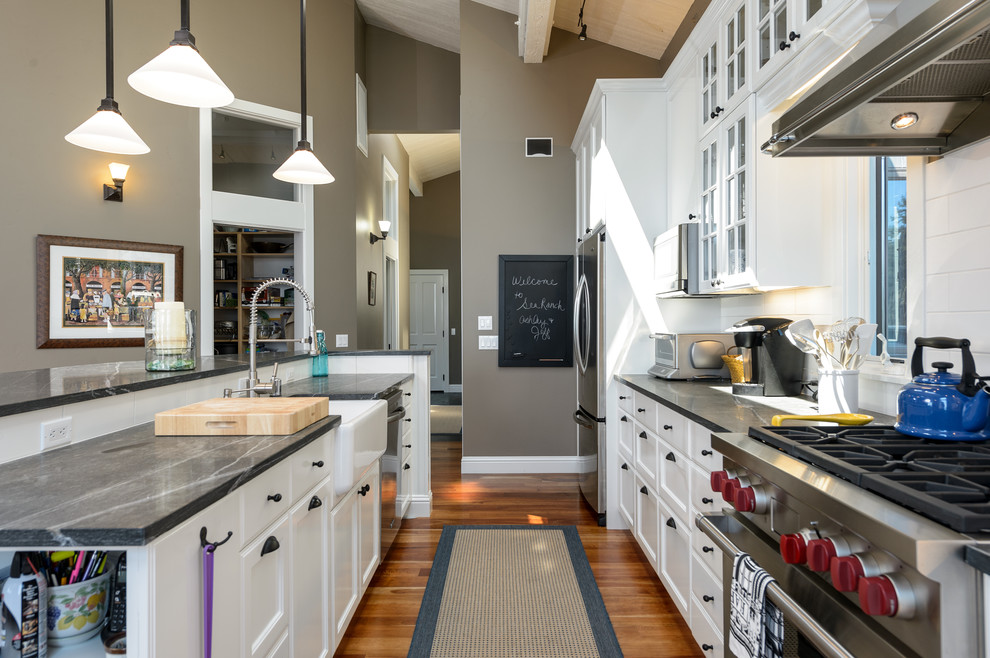 Photo of a mid-sized beach style single-wall open plan kitchen in San Francisco with a farmhouse sink, shaker cabinets, white cabinets, soapstone benchtops, white splashback, subway tile splashback, stainless steel appliances, light hardwood floors, with island and brown floor.