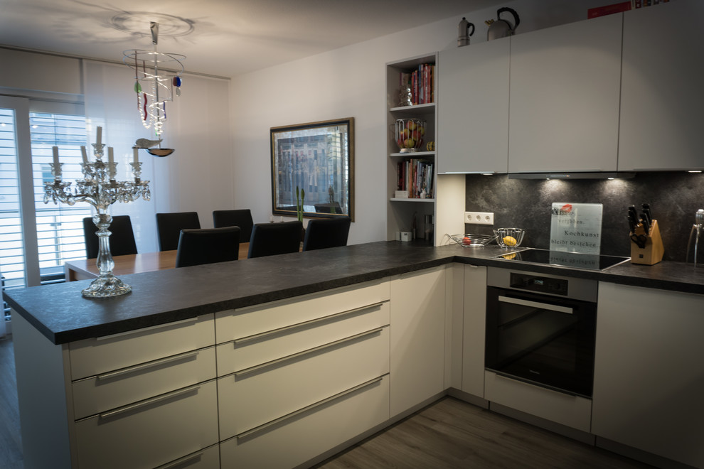 Photo of an expansive contemporary u-shaped open plan kitchen in Nuremberg with a drop-in sink, flat-panel cabinets, white cabinets, laminate benchtops, black splashback, timber splashback, black appliances, vinyl floors, a peninsula and black benchtop.