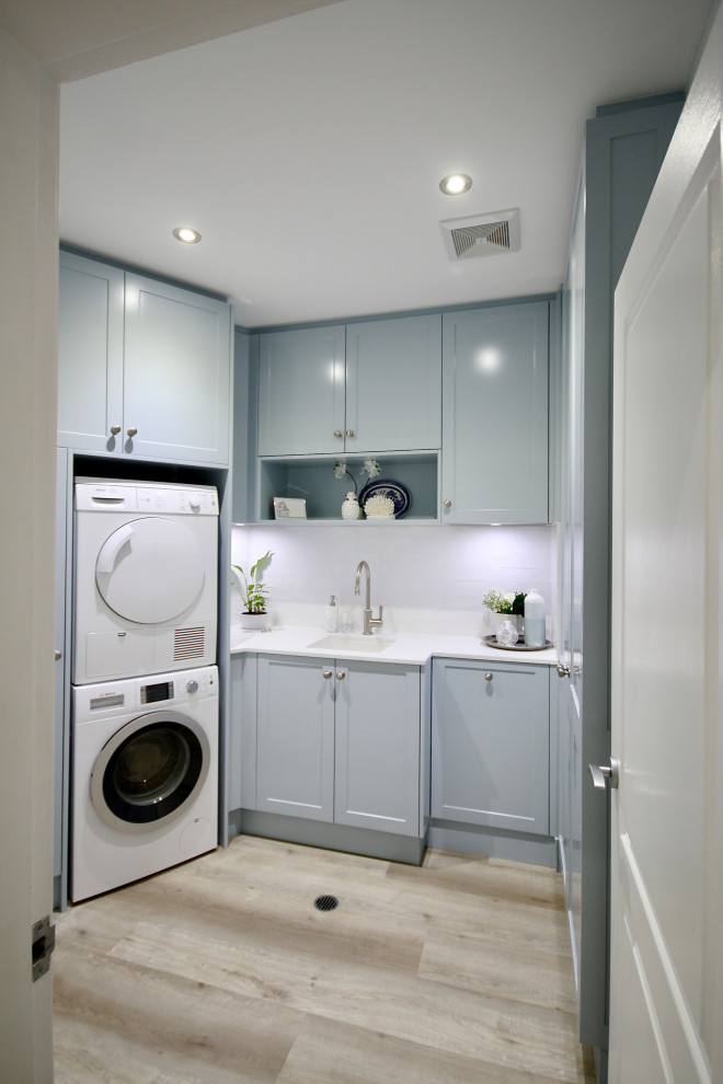 Inspiration for a mid-sized transitional u-shaped dedicated laundry room in Sydney with an undermount sink, shaker cabinets, blue cabinets, quartz benchtops, white splashback, subway tile splashback, white walls, light hardwood floors, a stacked washer and dryer, brown floor and white benchtop.