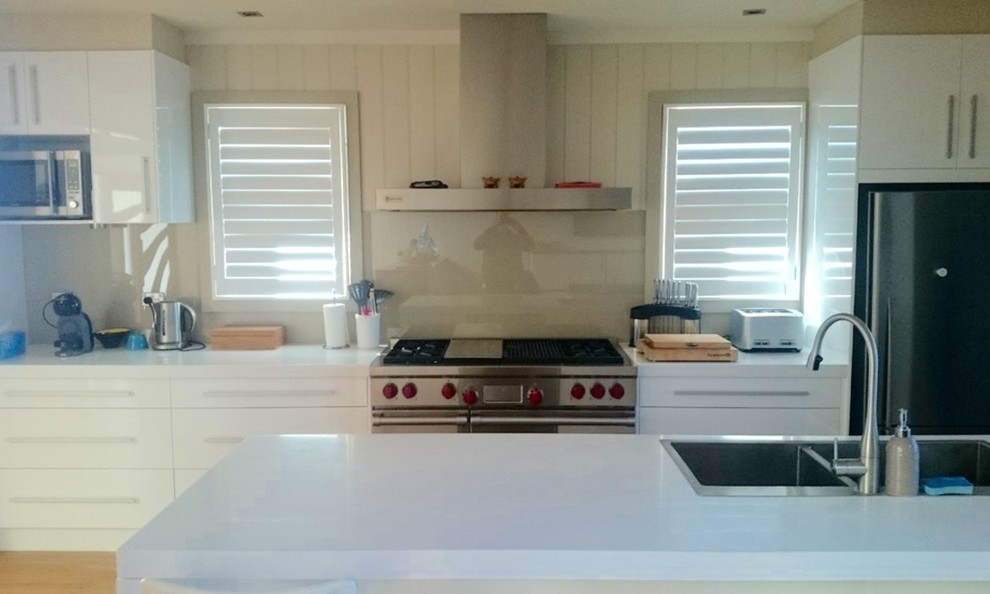 Photo of a mid-sized beach style single-wall open plan kitchen in Wollongong with a drop-in sink, flat-panel cabinets, white cabinets, quartz benchtops, white splashback, glass sheet splashback, stainless steel appliances, dark hardwood floors, with island, brown floor and white benchtop.