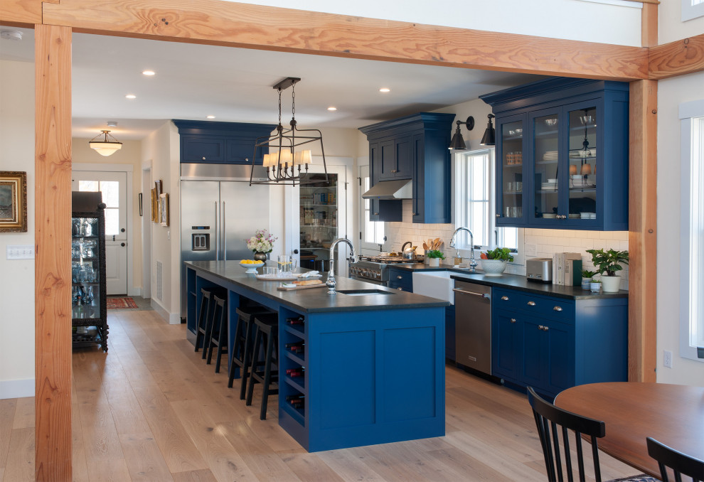 Photo of a mid-sized country single-wall eat-in kitchen in Boston with a farmhouse sink, shaker cabinets, blue cabinets, soapstone benchtops, white splashback, subway tile splashback, stainless steel appliances, light hardwood floors, with island and black benchtop.