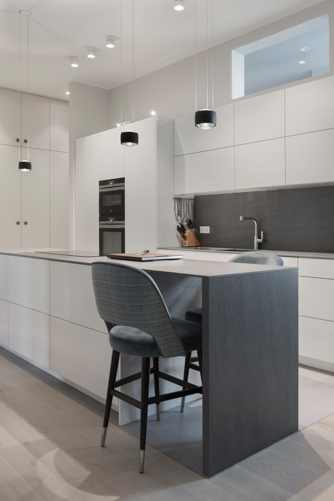 This is an example of a mid-sized contemporary kitchen in Berlin with flat-panel cabinets, white cabinets, grey splashback, black appliances, light hardwood floors, with island, beige floor and grey benchtop.