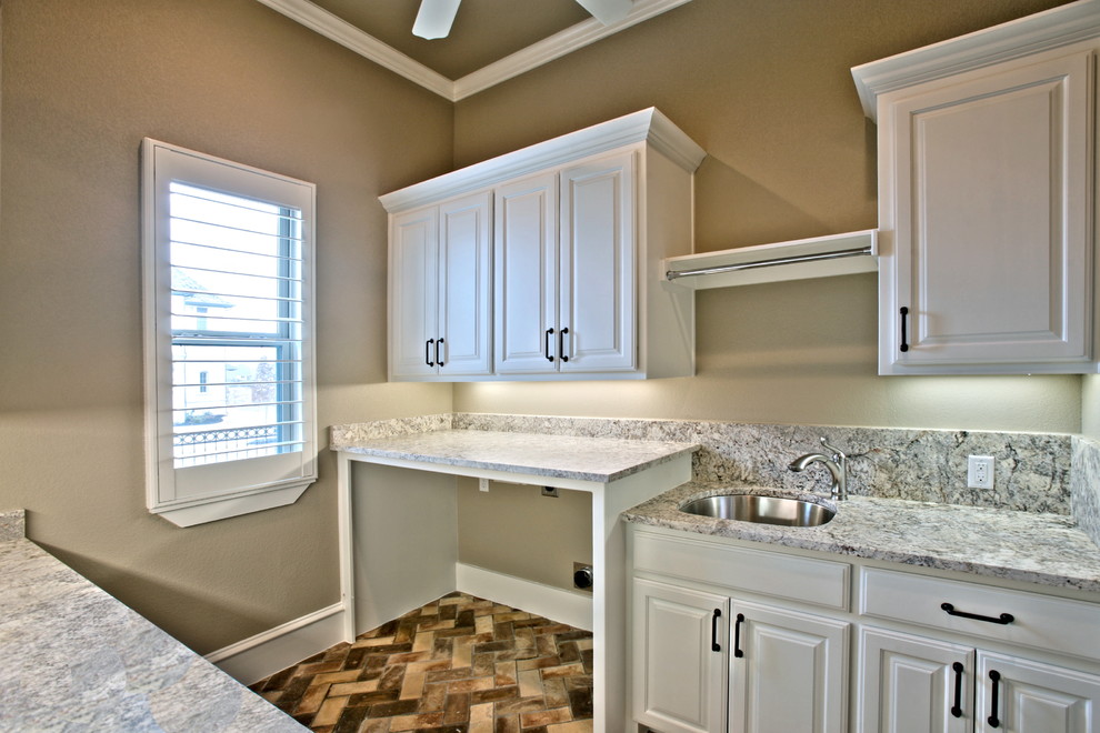 Mid-sized traditional galley dedicated laundry room in Dallas with a drop-in sink, shaker cabinets, white cabinets, granite benchtops, brown walls, brick floors and a side-by-side washer and dryer.