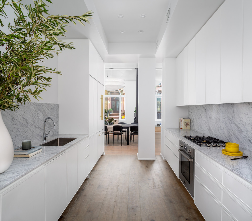 Inspiration for a large contemporary galley open plan kitchen in New York with a single-bowl sink, flat-panel cabinets, white cabinets, marble benchtops, white splashback, marble splashback, stainless steel appliances, light hardwood floors, no island, brown floor and white benchtop.