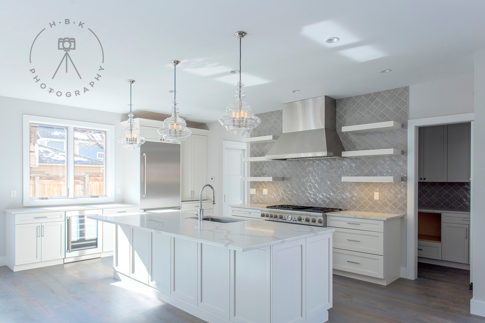 Example of a large single-wall medium tone wood floor and gray floor open concept kitchen design with a drop-in sink, shaker cabinets, white cabinets, marble countertops, gray backsplash, ceramic backsplash, stainless steel appliances, an island and white countertops