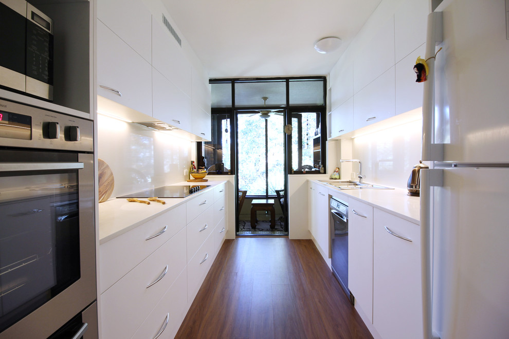 This is an example of a small modern galley separate kitchen in Sydney with a double-bowl sink, flat-panel cabinets, white cabinets, quartz benchtops, stone slab splashback, stainless steel appliances, vinyl floors, no island, brown floor, white splashback and white benchtop.
